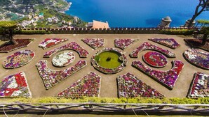 Ein Garten mit geometrischen Blumenbeeten und einem zentralen Teich, mit Blick auf eine Küstenlandschaft.