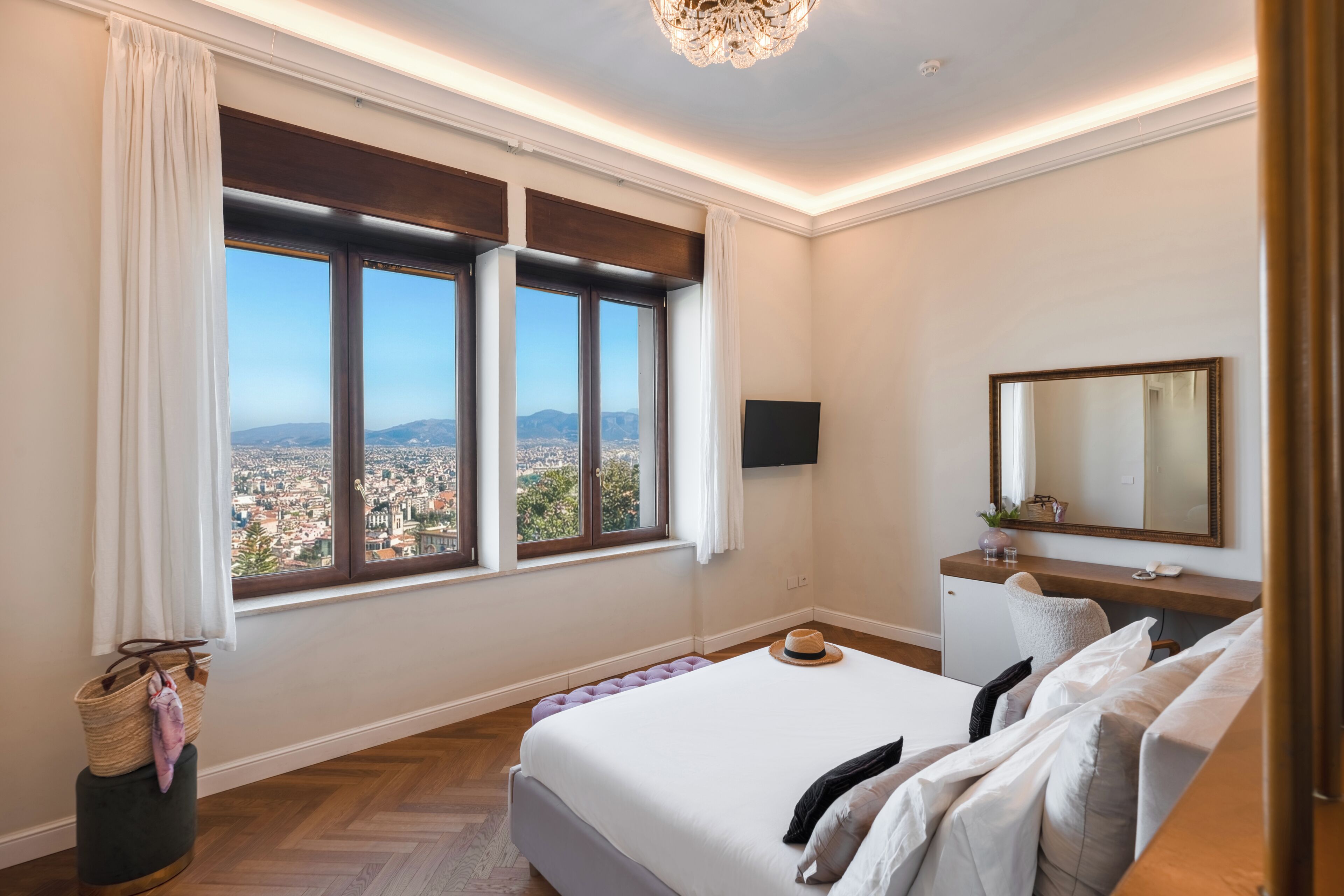 A bedroom with a large window, a bed, a bedside table, a mirror, and a view of a cityscape.