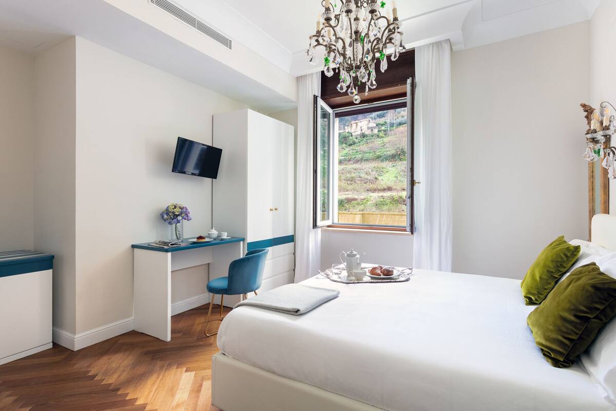 Ett hotellrum med en säng, ett skrivbord med en stol, en tv och ett fönster med utsikt över grönområden.