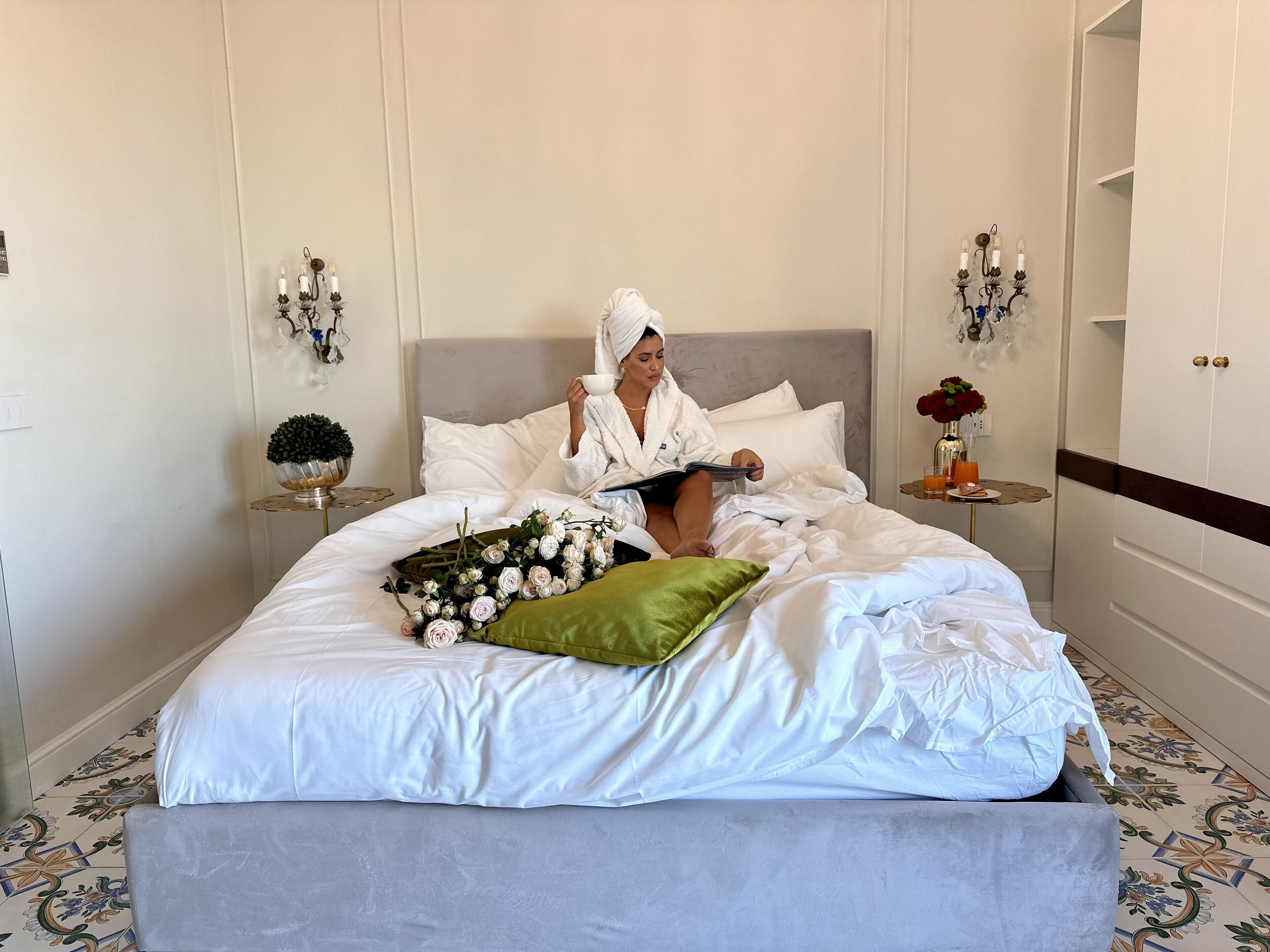 A neatly made bed with white linens, a green pillow, and a bouquet of flowers.