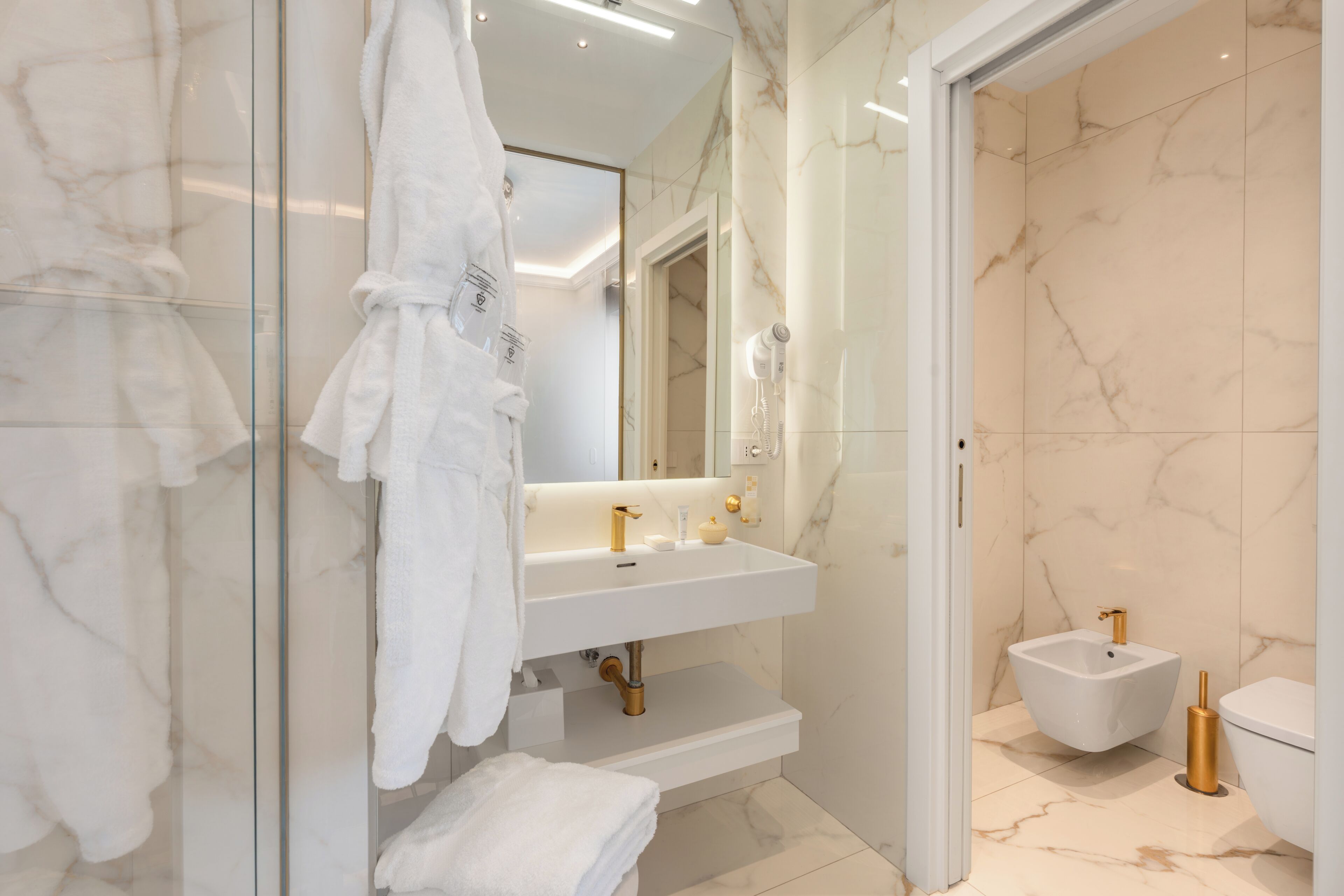 Un baño moderno con cabina de ducha de vidrio, un lavamanos blanco con detalles dorados, un bidé y una bata blanca colgada en la pared.