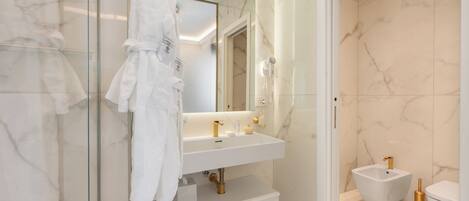 Un baño moderno con cabina de ducha de vidrio, un lavamanos blanco con detalles dorados, un bidé y una bata blanca colgada en la pared.