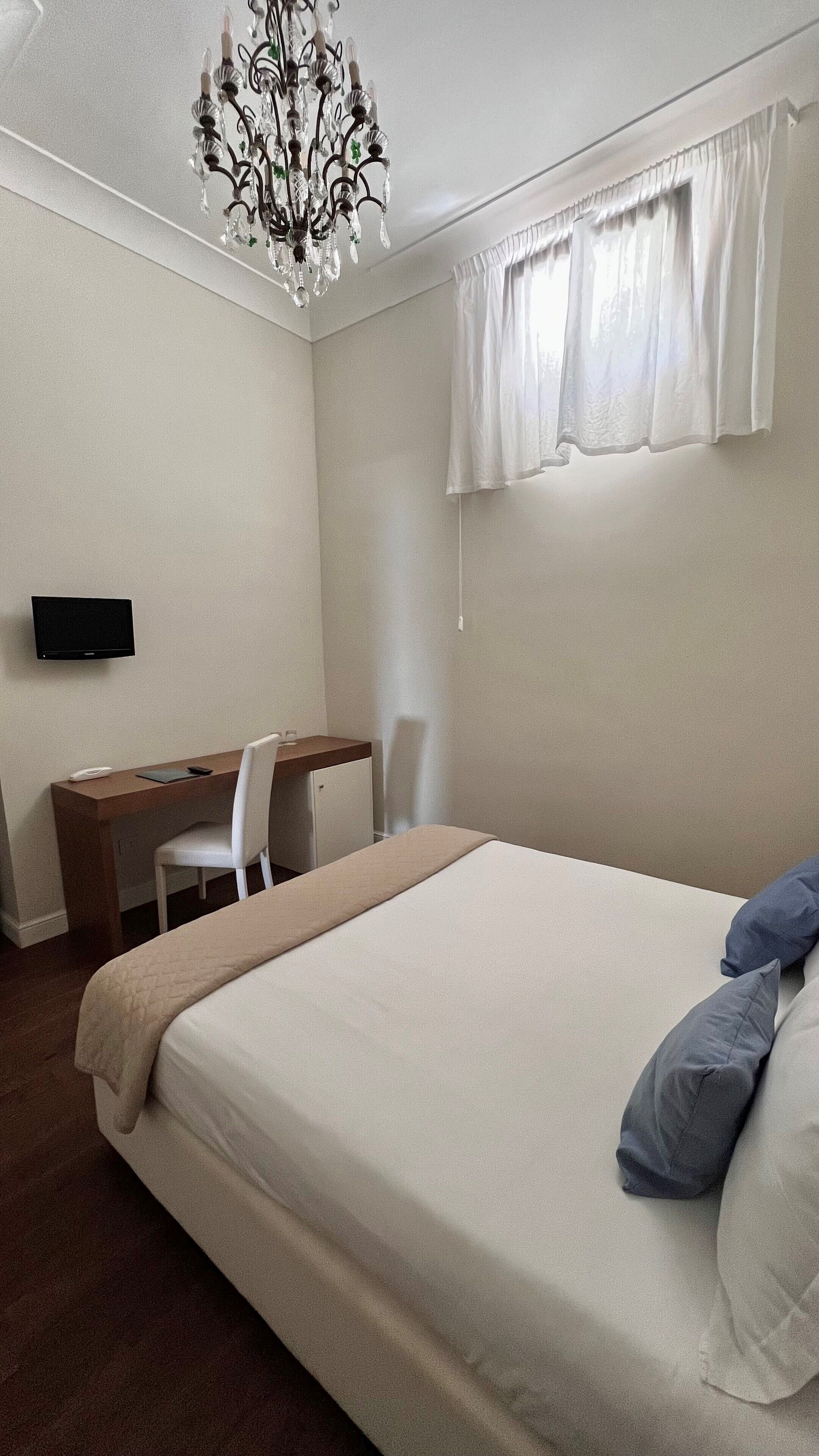 A bedroom with a bed, a desk, a chair, a television, and a chandelier.