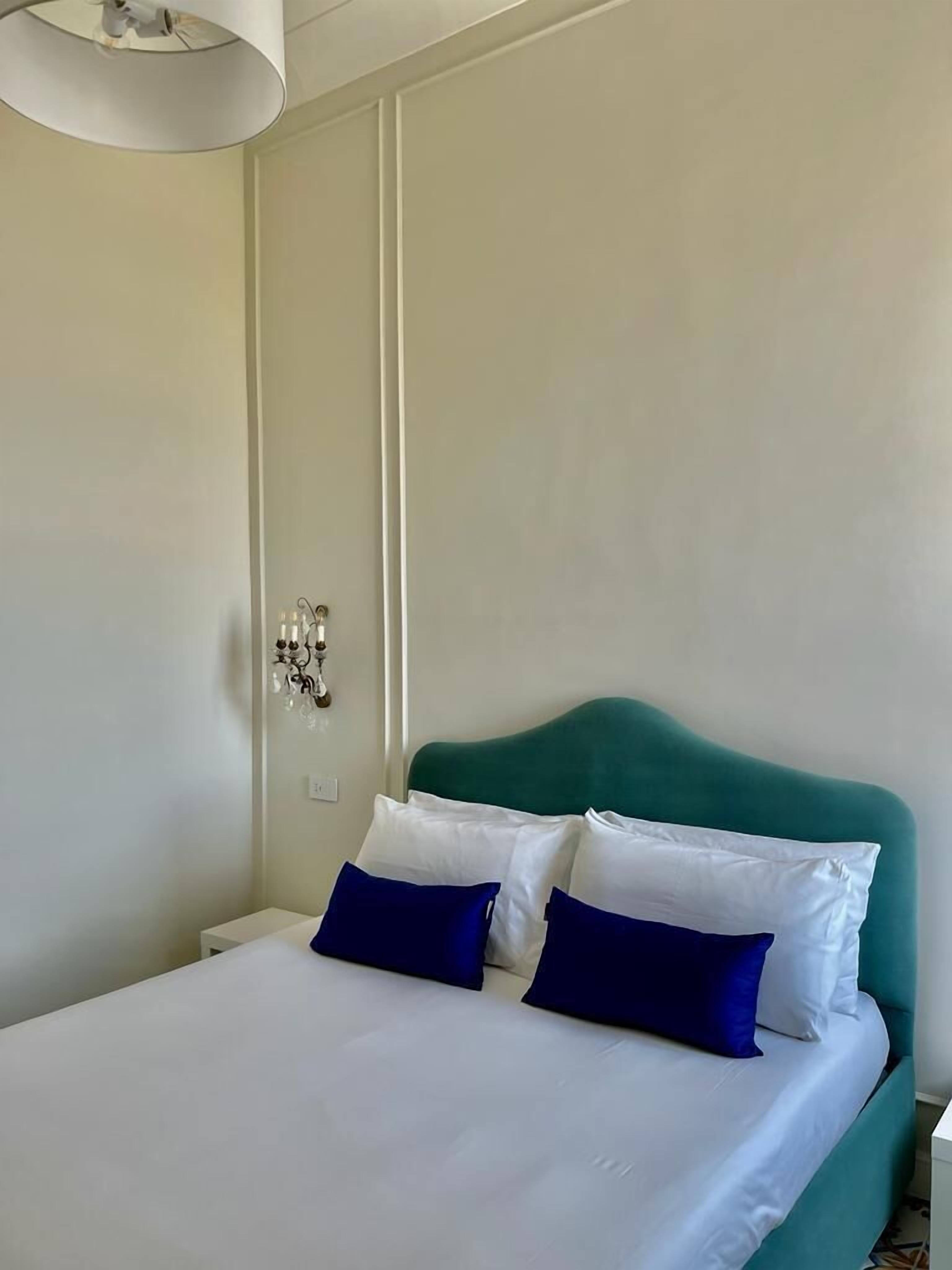 Een net opgemaakt bed met witte en blauwe kussens, een blauwgroen hoofdbord en een witte nachtkastje met een lamp.