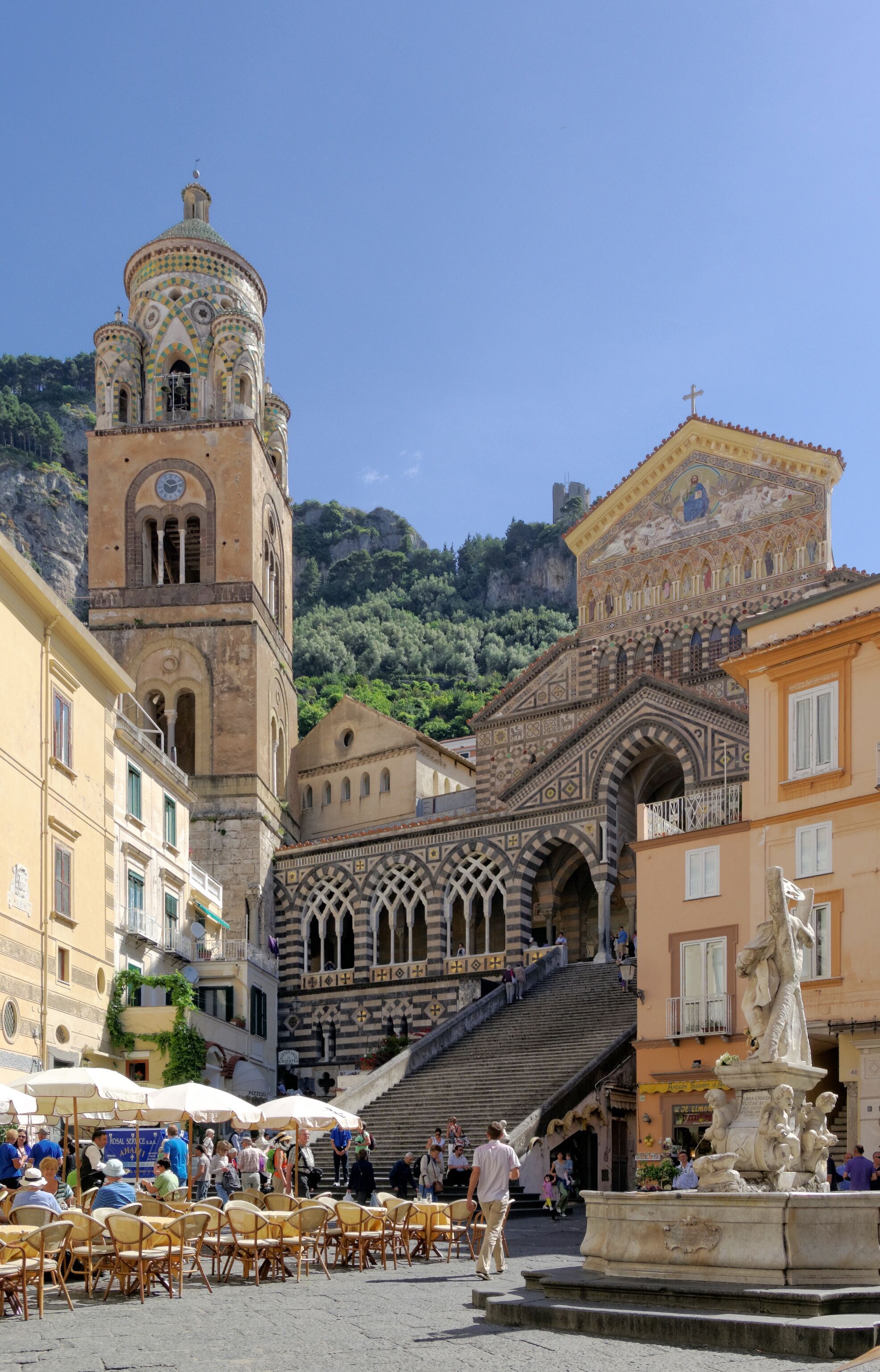 Une place historique, dominée par le clocher d’une église imposante, agrémentée d’une fontaine et d’un escalier menant à un bâtiment à l’architecture complexe.