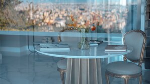 Ein runder Tisch mit Blumen, zwei Bücher und ein Stuhl in einem Raum mit Blick auf eine Stadtlandschaft durch ein Fenster.