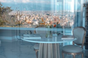Una mesa redonda con un florero, dos libros y una silla en una habitación con vistas a un paisaje urbano a través de un ventanal.