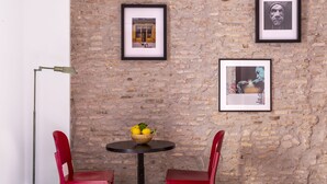 Una habitación con pared de ladrillo, dos sillas rojas, una mesa redonda con un frutero y dos cuadros enmarcados.