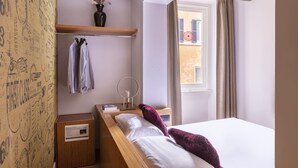Un dormitorio con cama, mesita de noche, espejo, estante con ropa y ventana con cortinas.