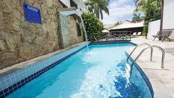 A swimming pool with a stone wall and a sign, surrounded by palm trees and a covered area with lounge chairs.
