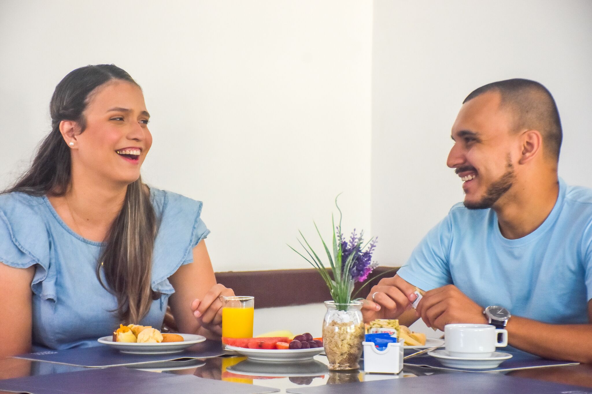 Two people sitting at a table with breakfast items, including fruits, a glass of orange juice, and a small vase with flowers.