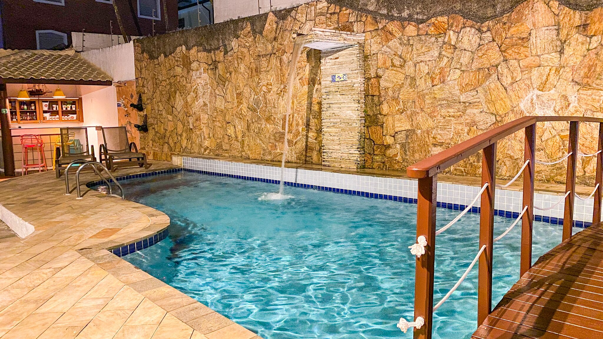 A swimming pool with a stone wall backdrop, a wooden deck, and a bar area with chairs.