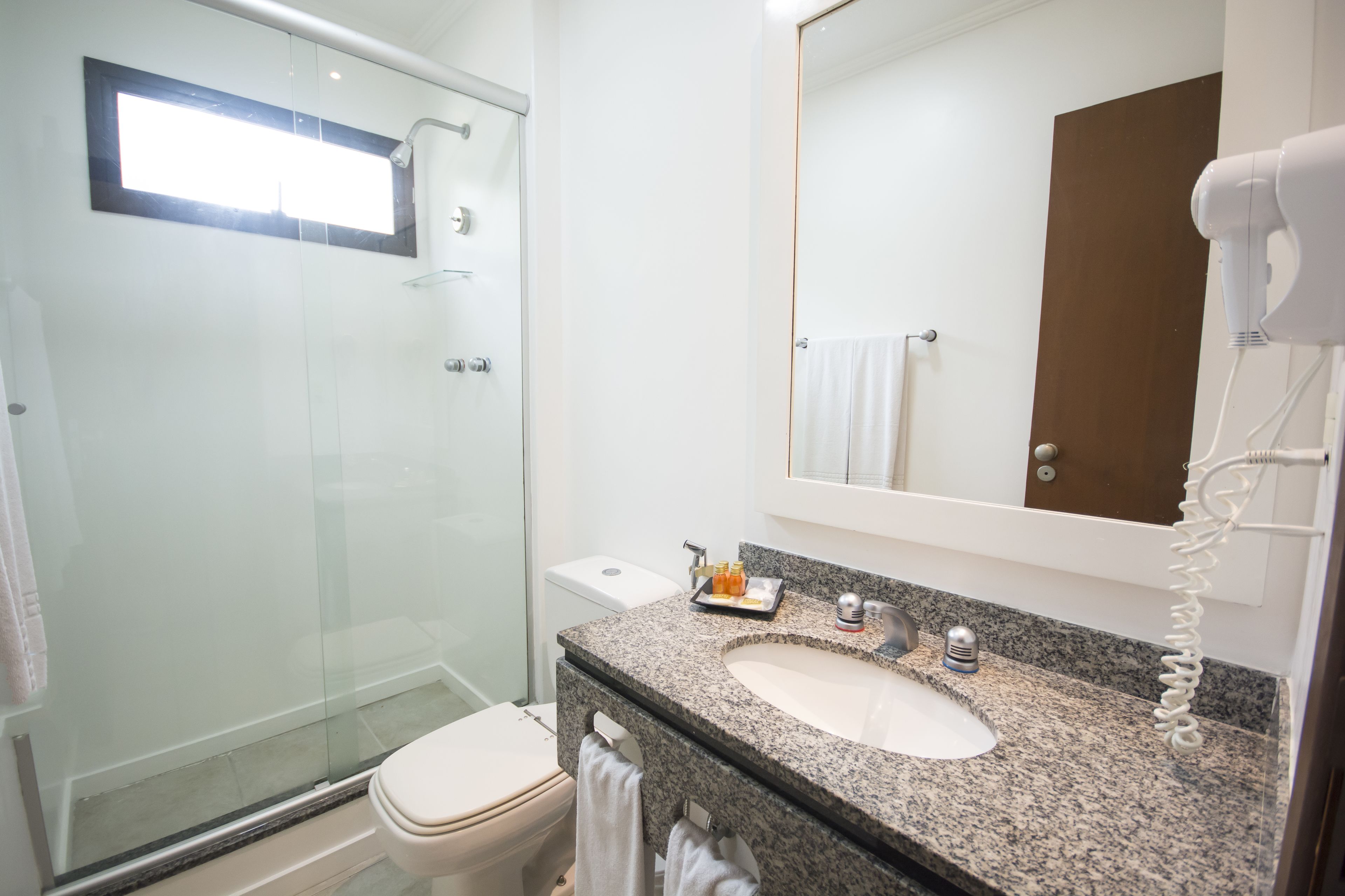 Une salle de bain moderne avec une douche à parois de verre, des toilettes blanches, un lavabo en granit et un sèche-cheveux posé sur le comptoir.