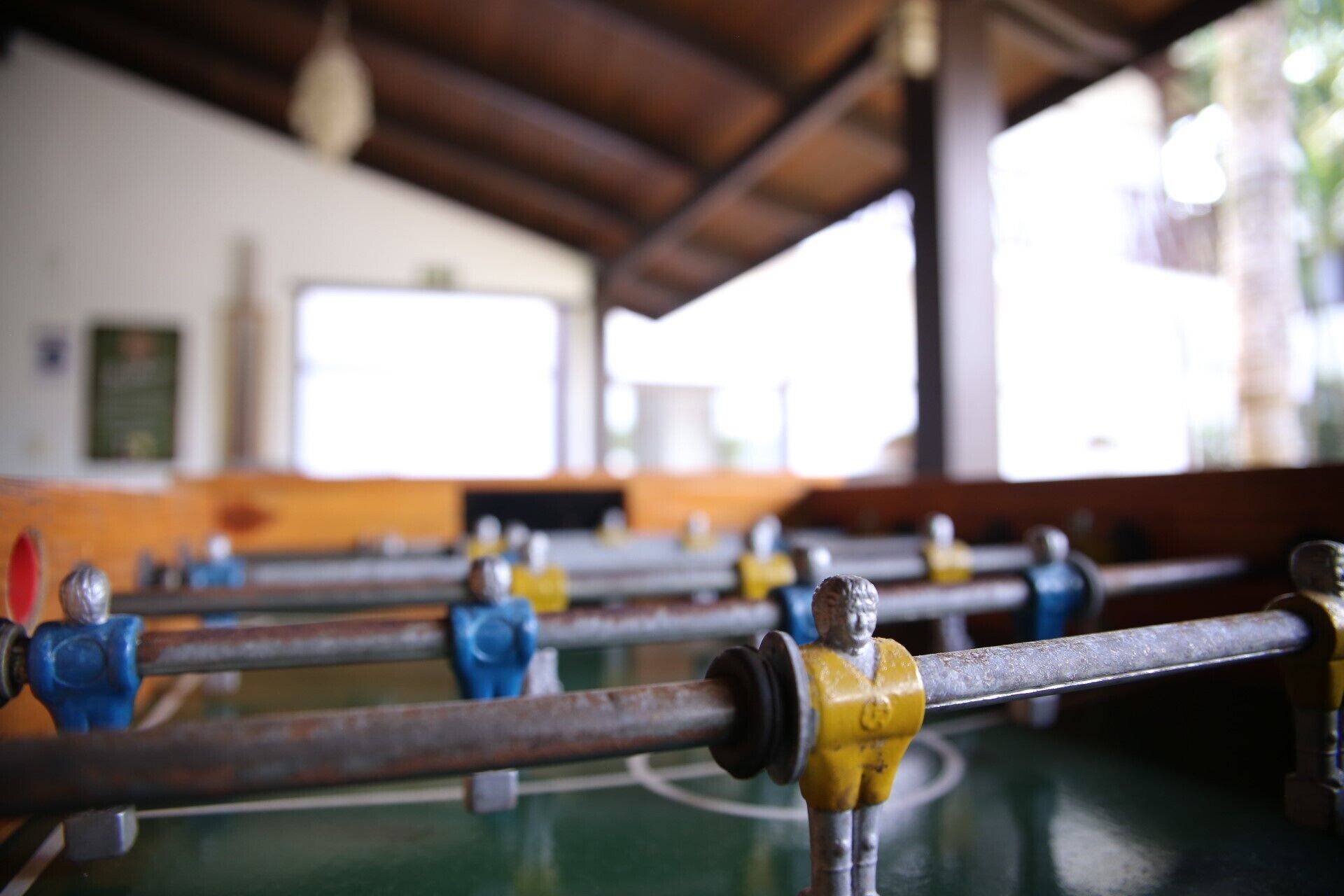 A foosball table with metal rods and figures, set in a room with a wooden ceiling and large windows.