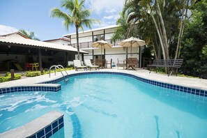 A swimming pool with lounge chairs and umbrellas, surrounded by palm trees and a building with a tiled roof. - Hotel Ilhas da Grécia (Guarujá)