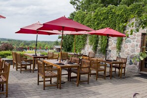 An outdoor patio with wooden tables and chairs, red umbrellas, and a stone wall with greenery.