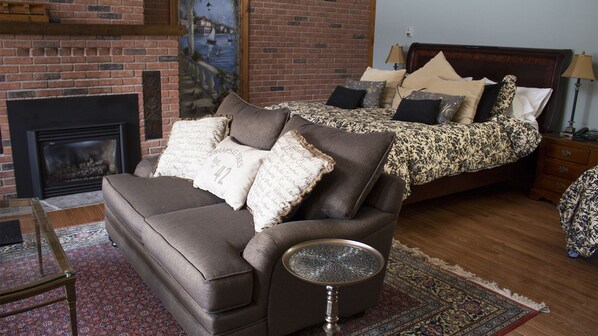 A bedroom with a sofa, a round side table, a bed with patterned bedding, a nightstand, a lamp, and a fireplace.