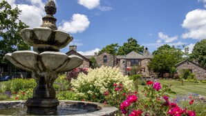 A stone fountain with a tiered design, surrounded by a garden with various flowers and a stone building in the background. - Ste. Anne's Spa (Alnwick/Haldimand)