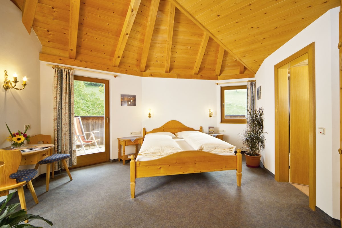A bedroom with a wooden bed, a desk with a chair, a potted plant, and a window with curtains.