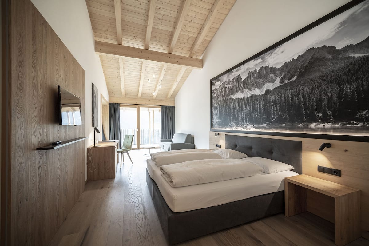 A modern bedroom with a large bed, a wooden headboard, a mounted TV, and a large mountain landscape painting.