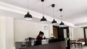 A reception area with a dark reception desk, multiple pendant lights, a clock on the wall, and a seating area with a bench and a table. - Suklutai Hotel & Serviced Apartment (Bang Khla)