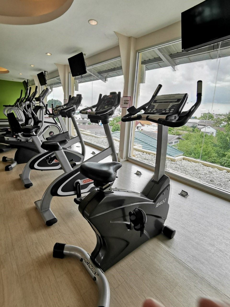 Une rangée de vélos d’appartement dans une salle de sport dotée de grandes fenêtres donnant sur un quartier résidentiel.