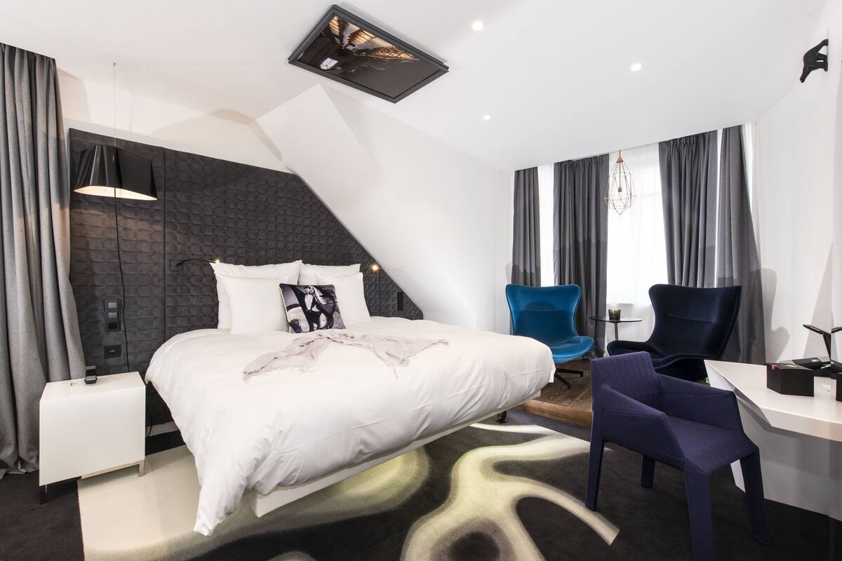 A modern hotel room with a large bed, a desk, a chair, and a unique ceiling design.