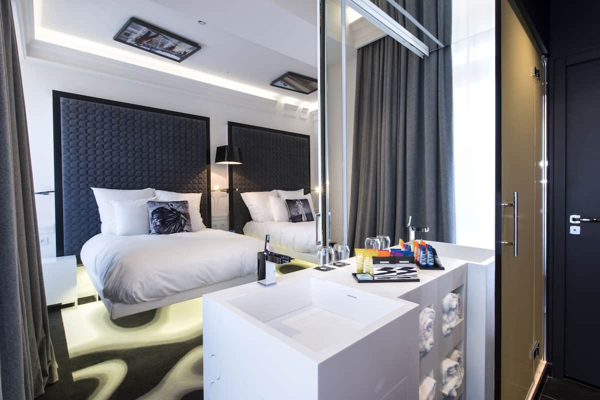 A modern hotel room with a large bed, a white bedside table, a built-in sink, and a large mirror.