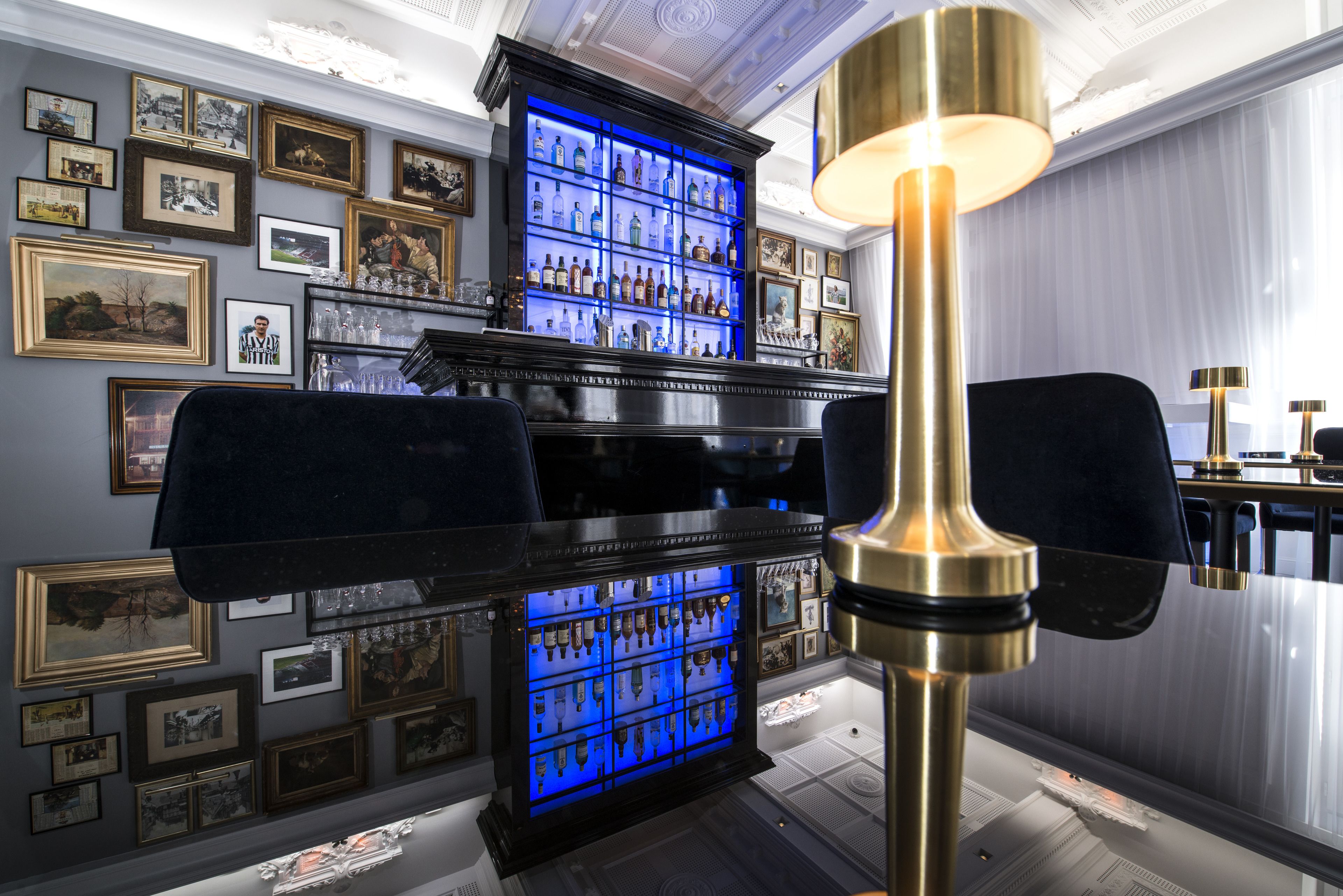 Un espace bar avec un présentoir illuminé de lumière bleue où sont exposées les bouteilles, des tableaux encadrés accrochés au mur et une grande lampe décorative.