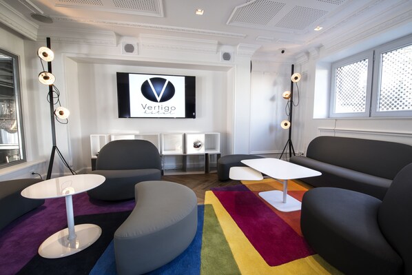 A modern hotel room with a large TV mounted on the wall, a comfortable seating area with a sofa and chairs, a small white table, and a colorful striped rug.