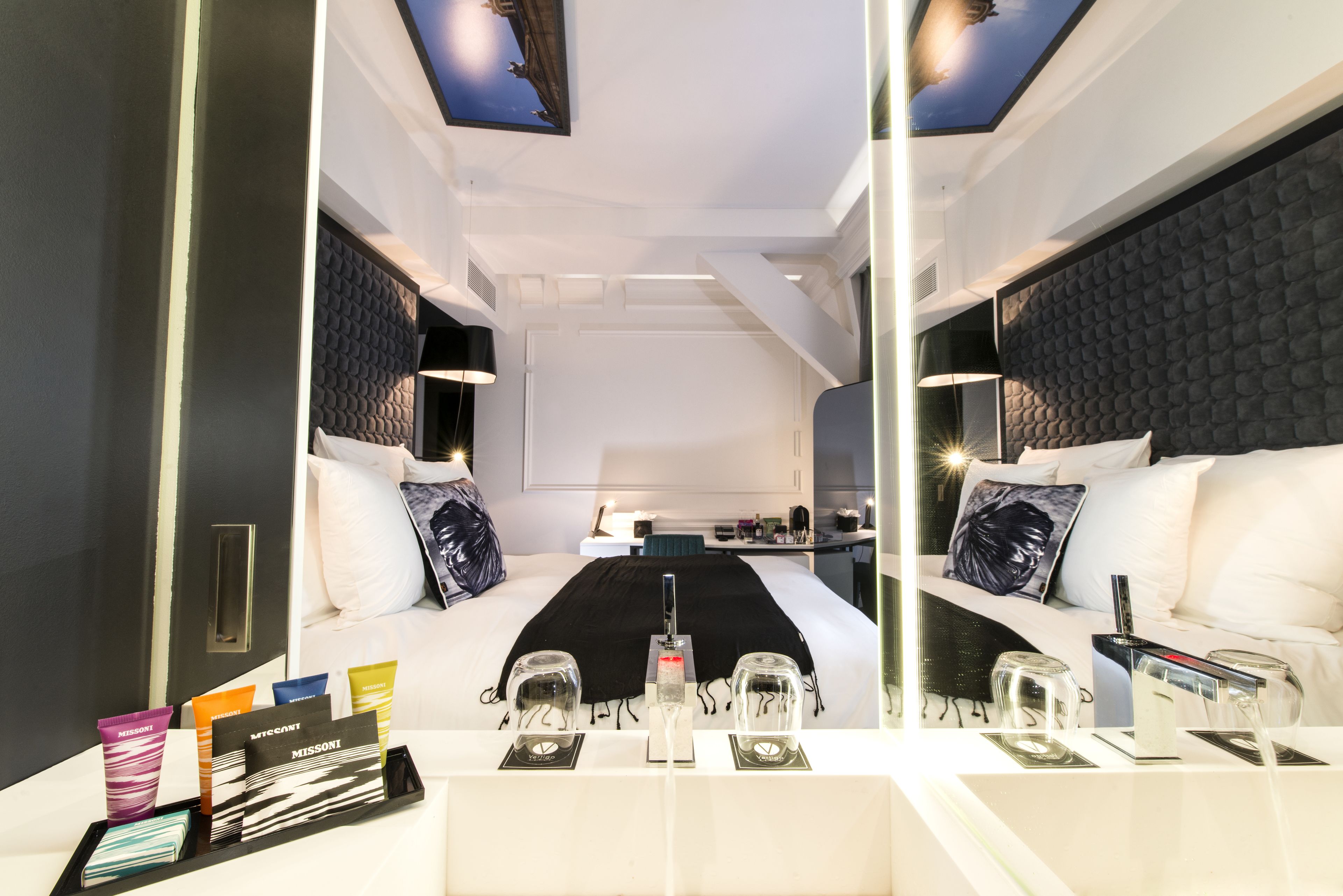 A modern hotel room with a large bed, bedside tables, a mirror, and a view of the sky through a skylight.