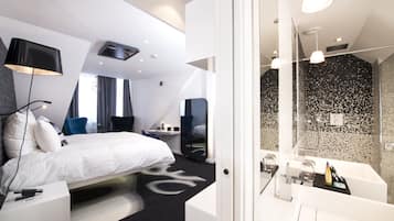 A modern hotel room with a large bed, a desk, a chair, a desk lamp, a mirror, a shower area, and a bathroom with a sink and mirror.