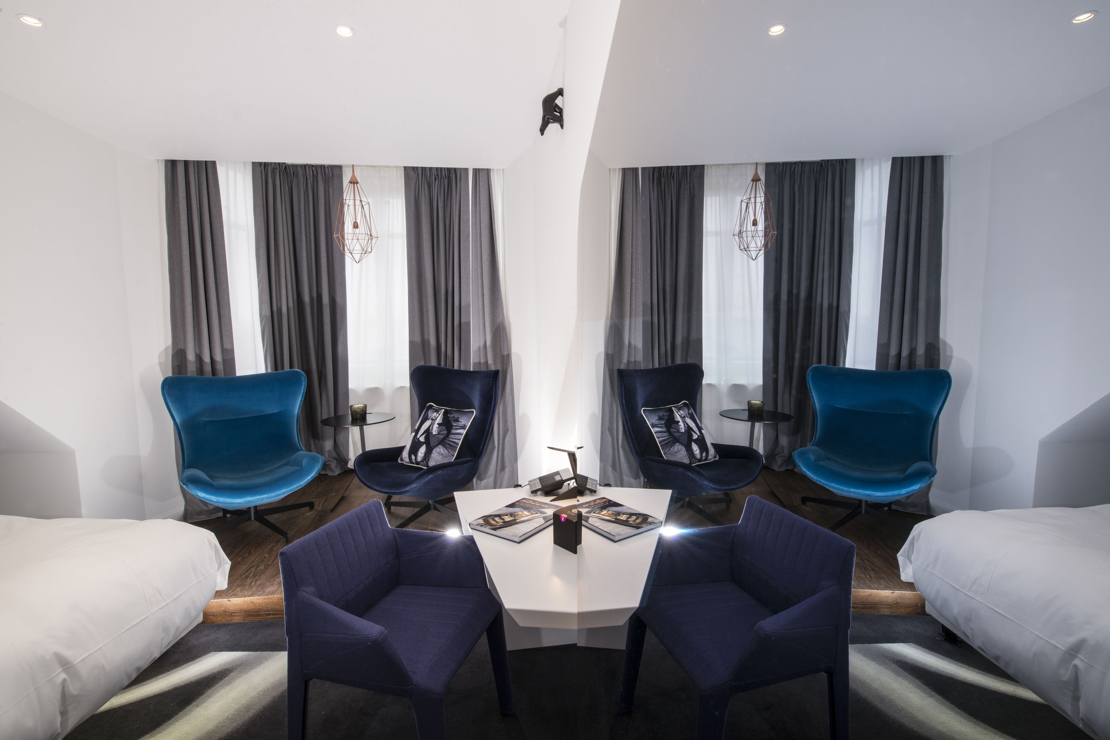 A modern hotel room with a central table, two blue chairs, a bed on each side, and a large window with grey curtains.