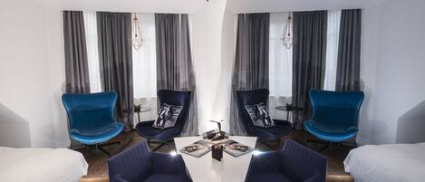 Habitación de hotel moderna con una mesa central, dos sillas azules, camas a ambos lados y un ventanal con cortinas grises.