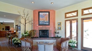 Un salon avec une cheminée, deux fauteuils en osier, une table basse et un grand tableau accroché au-dessus de la cheminée.