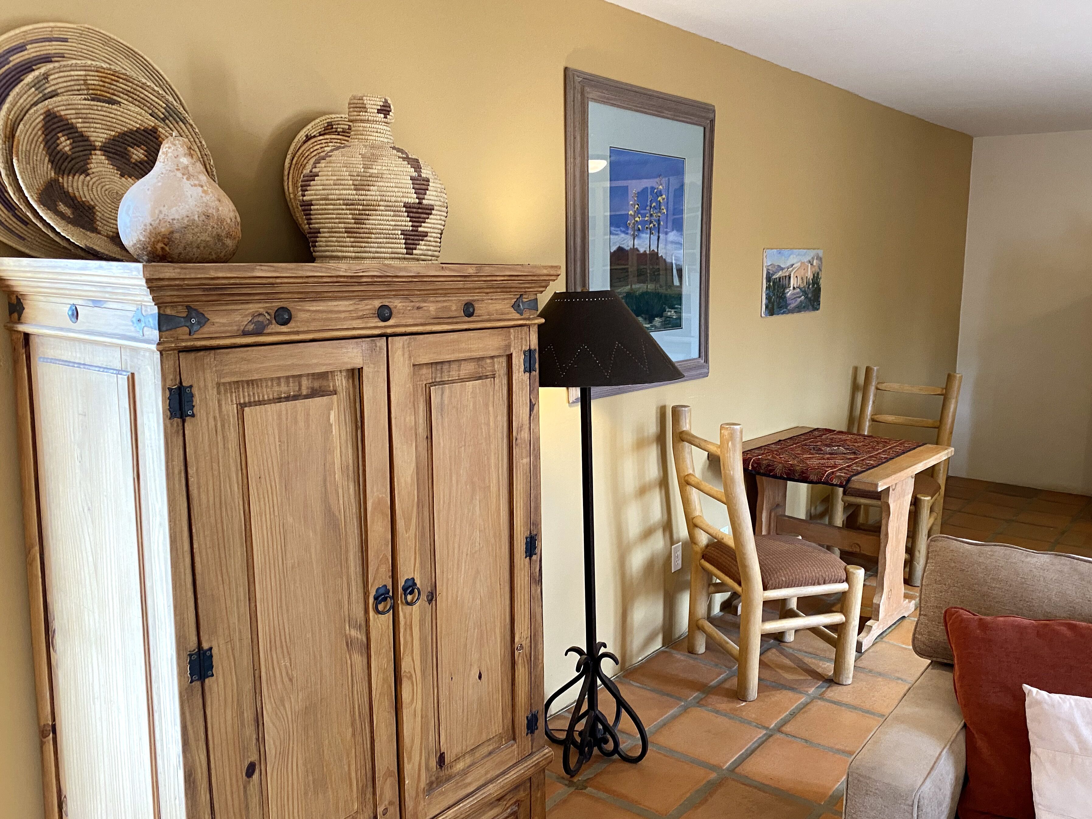 Un mueble de madera con pomos metálicos, un jarrón tejido, una lámpara, un cuadro enmarcado y una silla de madera con cojín.