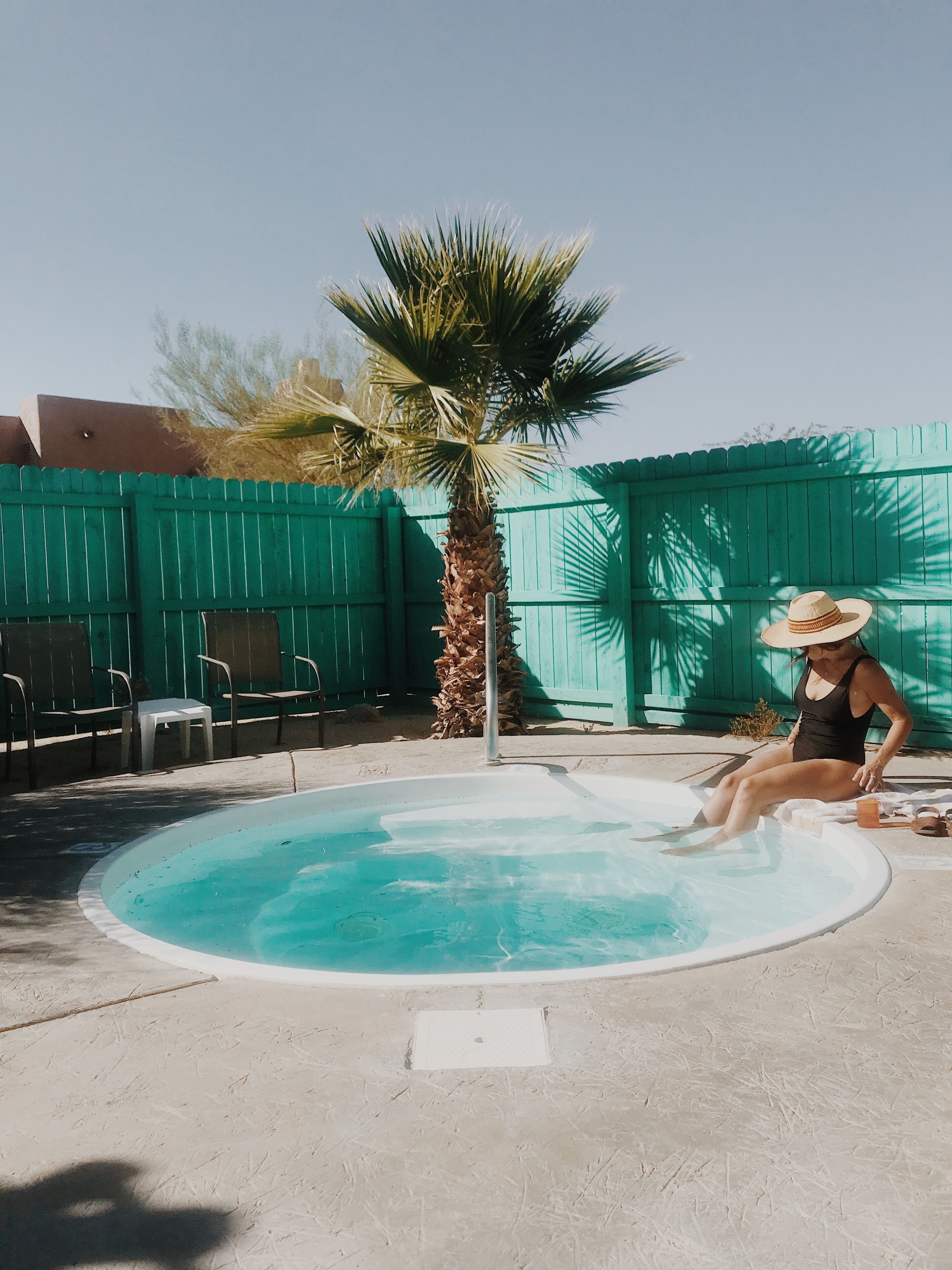 A person sitting by a small, round pool with a palm tree and a green fence in the background.