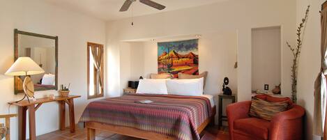 A bedroom with a bed, a chair, a desk, a ceiling fan, and a painting on the wall.