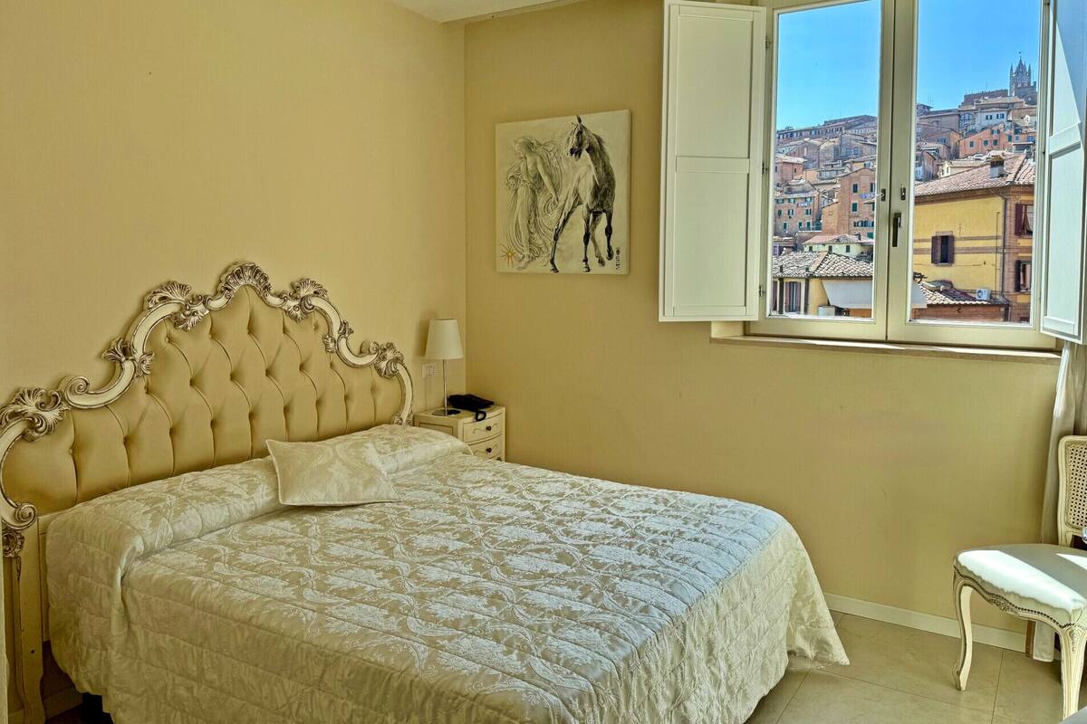 Un dormitorio con una cama grande, una mesita de noche, una silla, una ventana con vistas a la ciudad y un cuadro en la pared.
