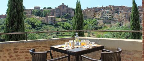 Un balcón con sillas de mimbre y una mesa preparada para el desayuno, con vista a un pueblo histórico de edificios escalonados y cipreses.
