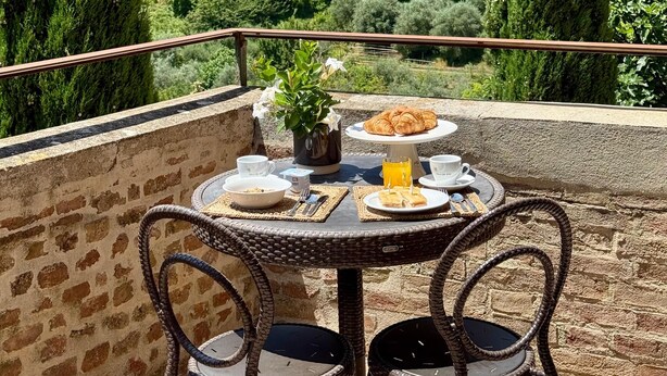Una terraza con sillas de mimbre y una mesa dispuesta para el desayuno, con vistas a un pueblo histórico de edificios en tonos terracota y cipreses.