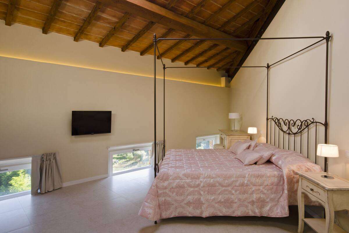 Un dormitorio con cama con dosel, un televisor de pantalla plana en la pared, mesitas de noche con lámparas y vistas a zonas verdes por las ventanas.