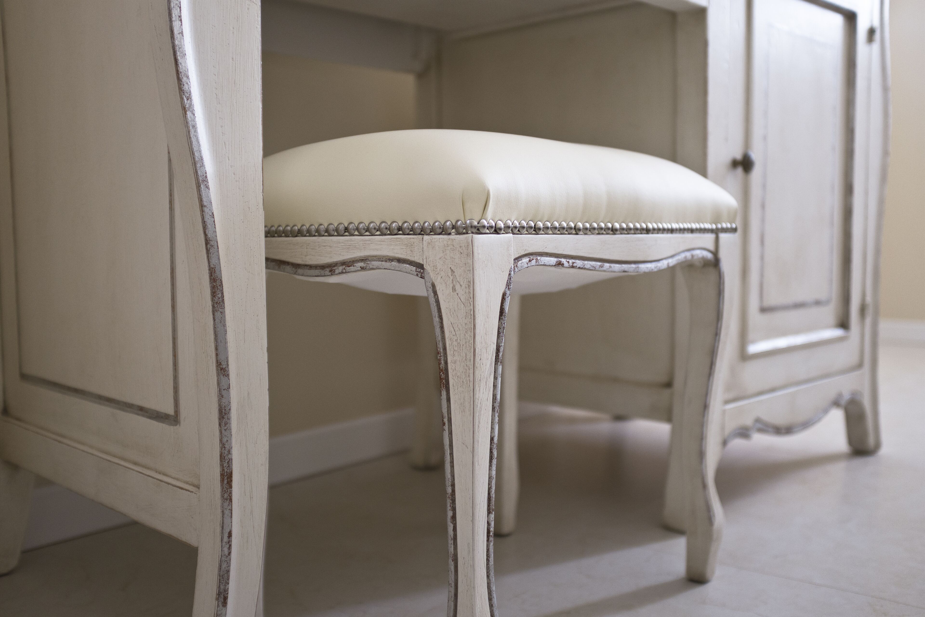 A white upholstered chair with a beaded edge, positioned in front of a white cabinet with a curved design.
