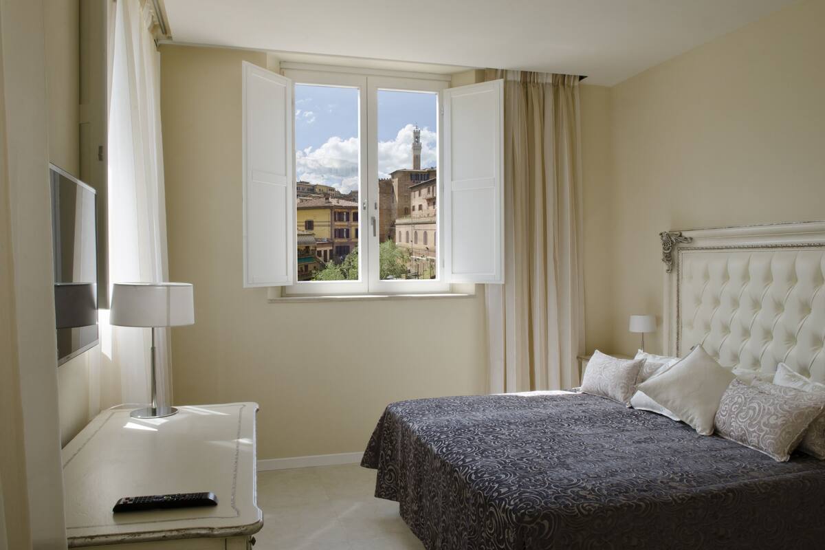 Una habitación de hotel con cama, televisión, ventana con cortinas y vistas a edificios.