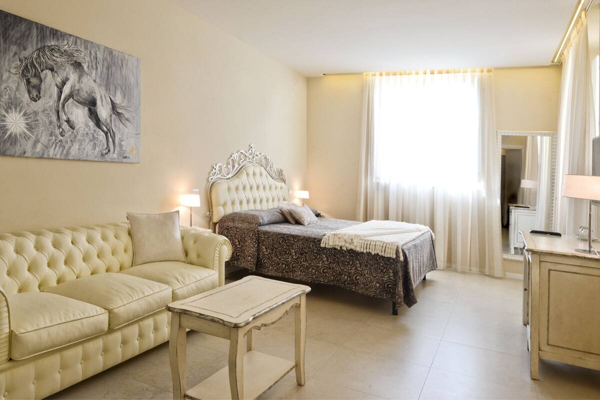 Una habitación de hotel con un sofá color crema con capitonado, una cama con colcha estampada, una mesita de noche, un ventanal con cortinas traslúcidas y un cuadro de un caballo en la pared.