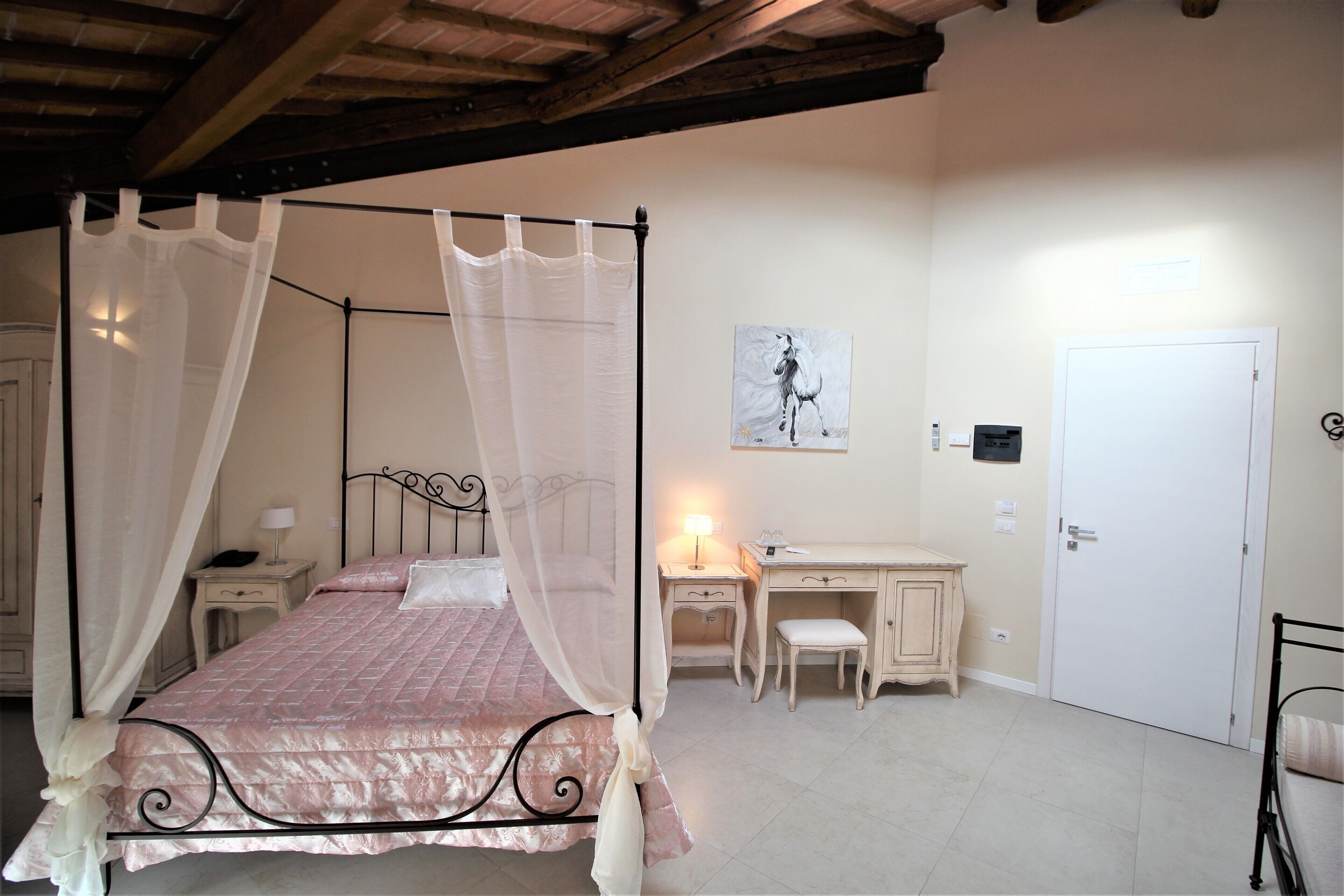 A bedroom with a four-poster bed, a desk, a chair, a nightstand, a lamp, and a door.