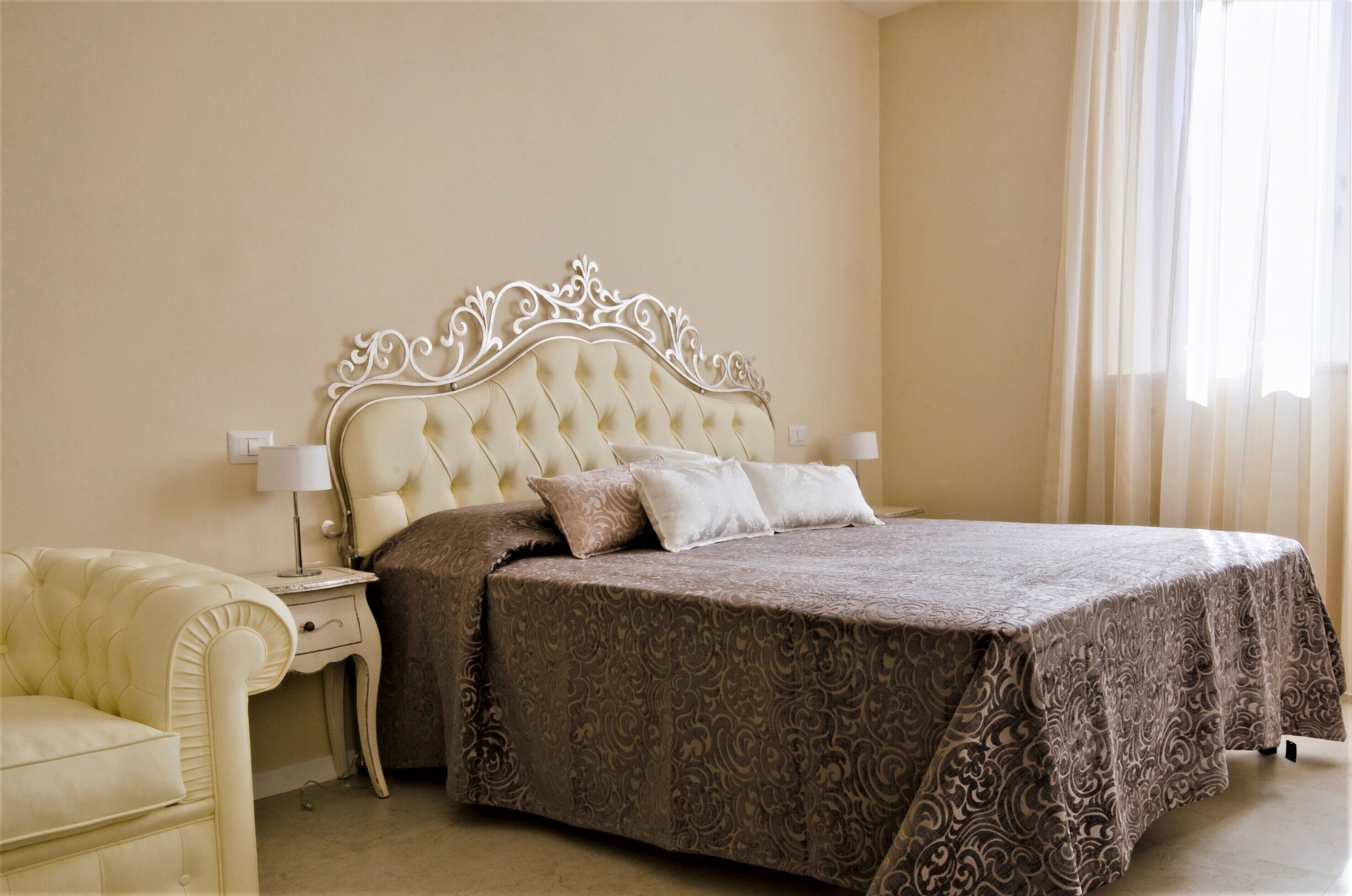 A bedroom with a tufted headboard, a large bed with a patterned bedspread, a bedside table with a lamp, and a chair.