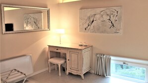 Una habitación con un escritorio blanco, una silla y un espejo grande. En la pared, hay un cuadro de un caballo y una ventana con cortinas.