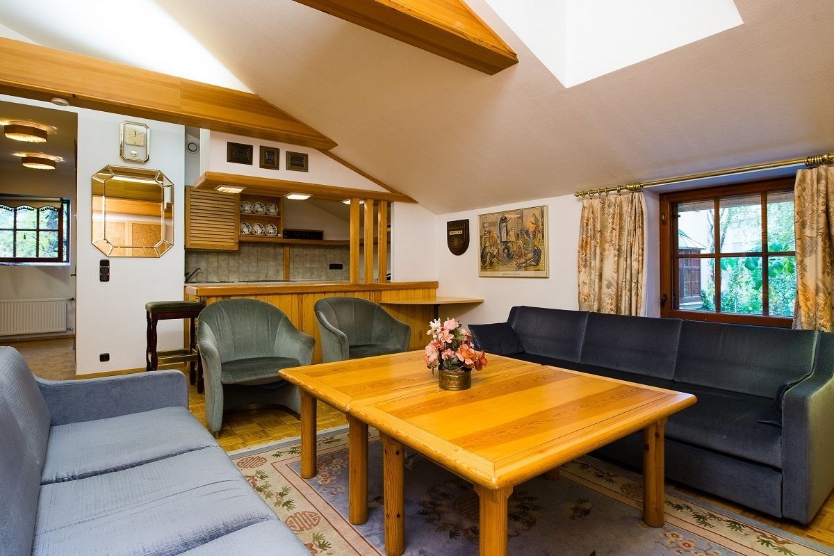 A cozy living room with a wooden table, a sofa, and a couch. There is a kitchen area with wooden cabinets and a window with curtains.