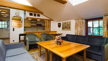 A cozy living room with a wooden table, a sofa, and a couch. There is a kitchen area with wooden cabinets and a window with curtains.